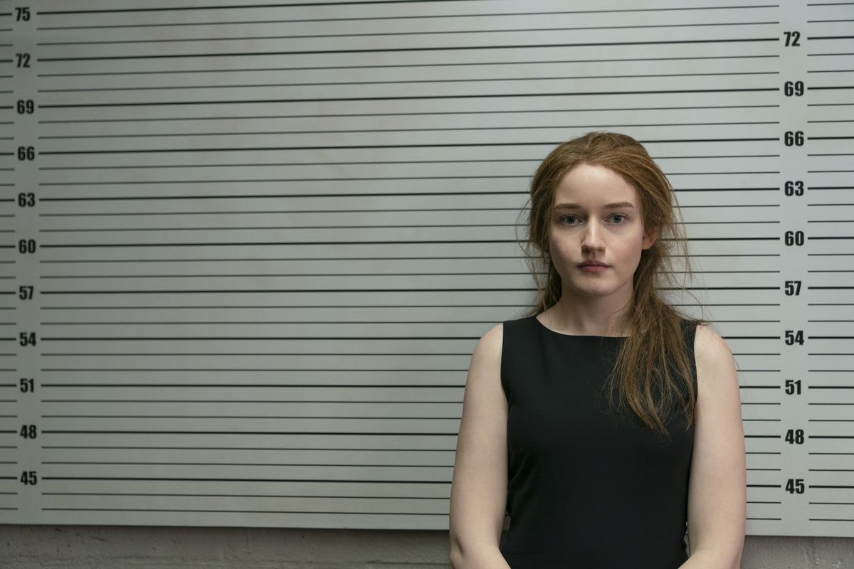 Inventing Anna: Julia Garner as Anna Delvey standing in front of a police lineup measuring thing